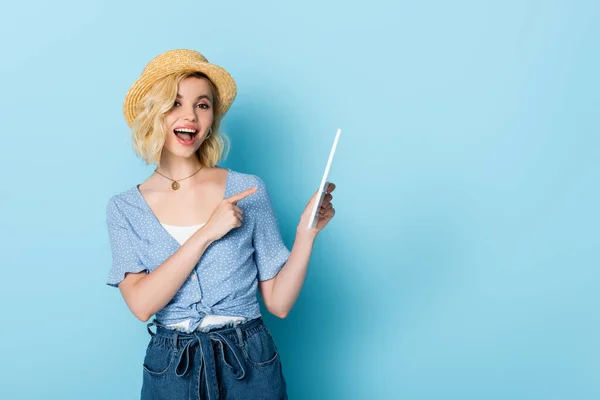 Femme excitée en chapeau de paille et shorts pointant du doigt la tablette numérique sur bleu — Photo de stock