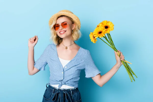 Молода жінка в солом'яному капелюсі і сонцезахисних окулярах тримає квіти на синьому — стокове фото