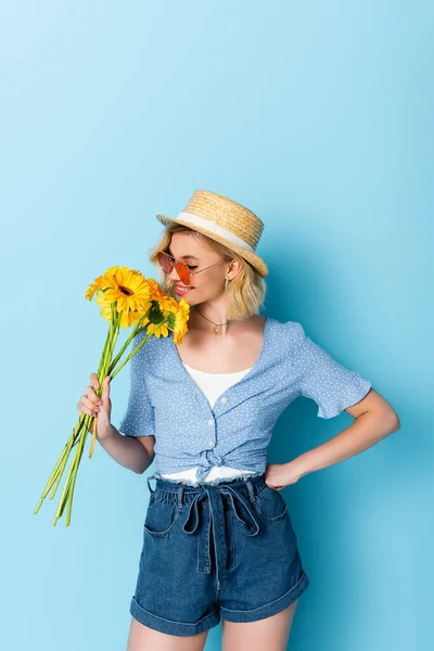 Young woman in straw hat and sunglasses smelling yellow flowers while standing with hand on hip on blue — Stock Photo