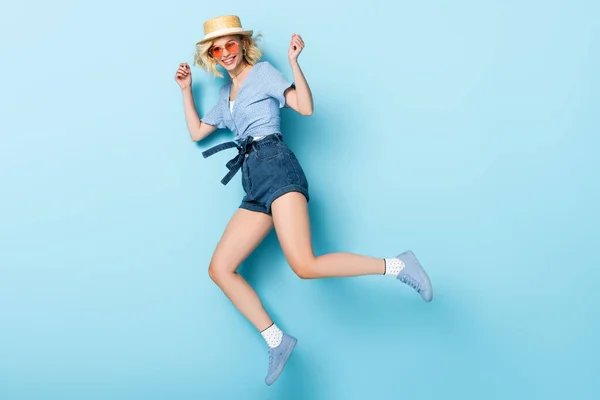 Жінка в солом'яному капелюсі і сонцезахисних окулярах стрибає на синьому — стокове фото