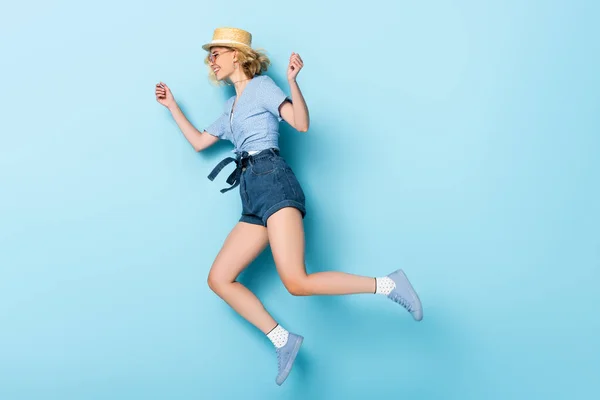 Вид сбоку на молодую женщину в соломенной шляпе и солнцезащитных очках, прыгающую на голубом — стоковое фото