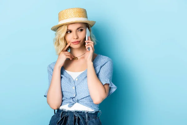 Mujer en sombrero de paja mirando hacia otro lado y hablando en el teléfono inteligente en azul - foto de stock