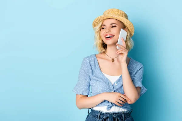 Женщина в соломенной шляпе с открытым ртом разговаривает на смартфоне на голубом — стоковое фото