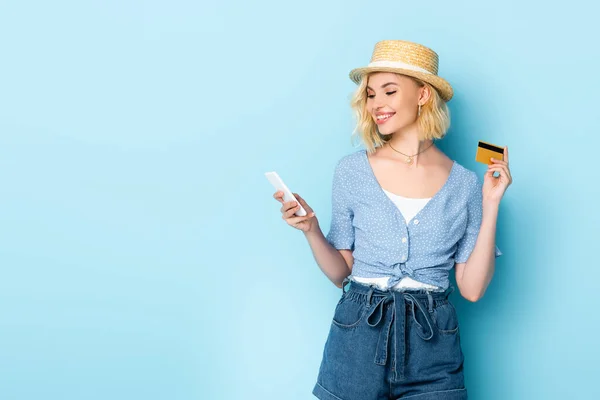 Mulher de chapéu de palha segurando cartão de crédito e smartphone em azul — Fotografia de Stock