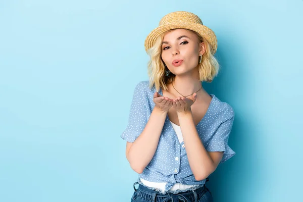 Joven mujer en paja sombrero enviando aire beso en azul - foto de stock