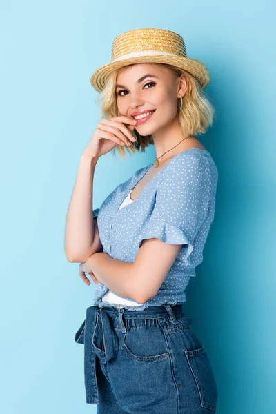 Giovane donna in cappello di paglia toccare il viso e guardando la fotocamera su blu — Foto stock