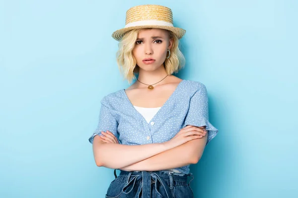 Ofendido joven mujer en sombrero de paja de pie con los brazos cruzados y mirando a la cámara en azul - foto de stock