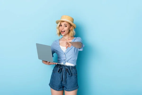 Foco seletivo da mulher em chapéu de palha segurando laptop e apontando com o dedo no azul — Fotografia de Stock