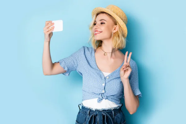 Mujer joven en sombrero de paja mostrando señal de paz y tomando selfie en azul - foto de stock