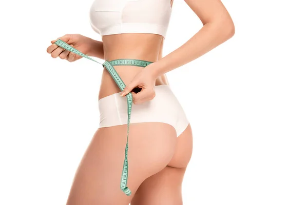 Vista recortada de una mujer joven sosteniendo cinta métrica mientras mide la cintura aislada en blanco - foto de stock