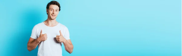 Imagen horizontal del hombre en camiseta blanca mostrando los pulgares hacia arriba sobre fondo azul - foto de stock