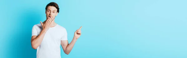 Panoramic shot of excited man in white t-shirt pointing with finger on blue background — Stock Photo