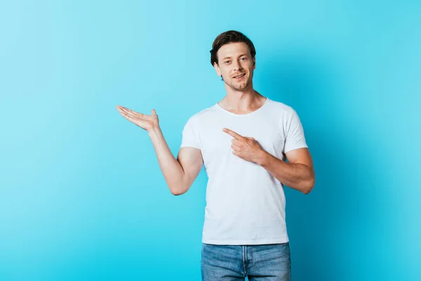 Mann im weißen T-Shirt zeigt mit Hand und Finger auf blauem Hintergrund — Stockfoto