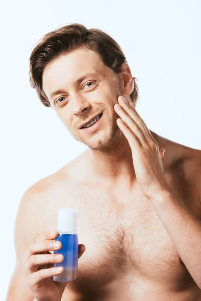 Uomo senza camicia toccando guancia e tenendo bottiglia di lozione isolata su bianco — Foto stock