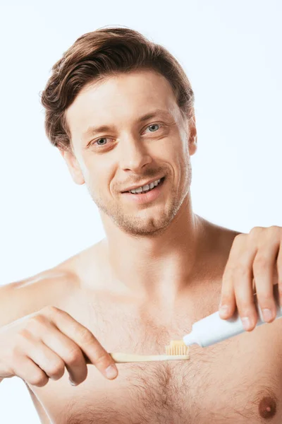 Селективный фокус человека без рубашки, смотрящего в камеру, держа зубную пасту и зубную щетку изолированными на белом — стоковое фото
