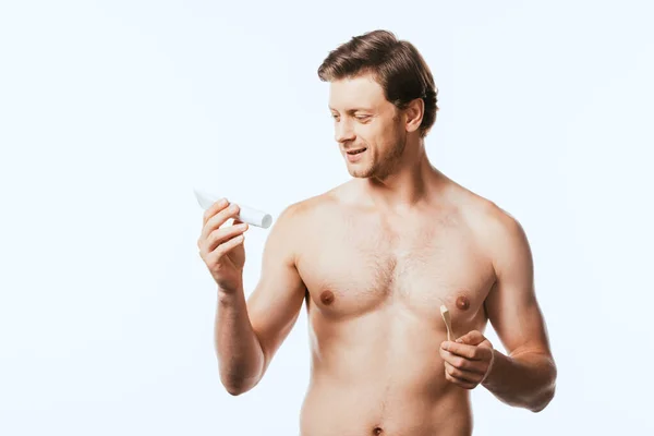 Hombre sin camisa sosteniendo el cepillo de dientes mientras mira la pasta de dientes aislada en blanco - foto de stock