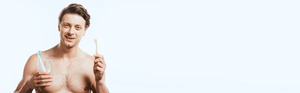 Panoramic shot of shirtless man holding glass with toothpaste and toothbrushes isolated on white — Stock Photo