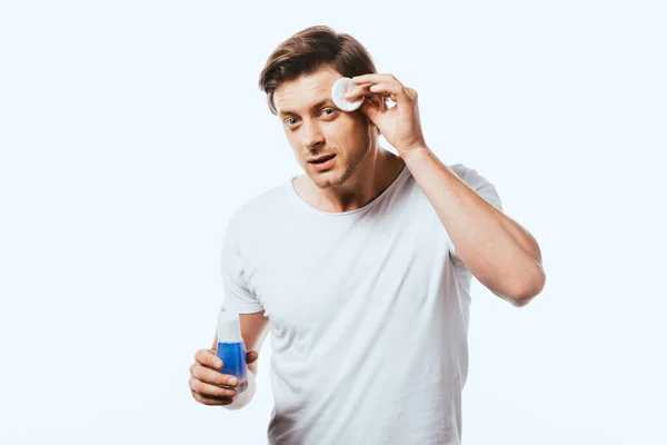 Joven mirando a la cámara mientras aplica loción con almohadilla de algodón aislada en blanco — Stock Photo