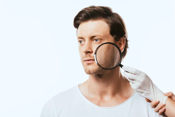 Jeune homme regardant loin tandis que dermatologue en gant de latex tenant loupe isolé sur blanc — Photo de stock