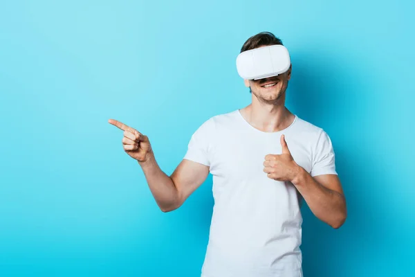 Hombre en camiseta blanca y auriculares vr mostrando el pulgar hacia arriba y señalando con el dedo sobre fondo azul - foto de stock