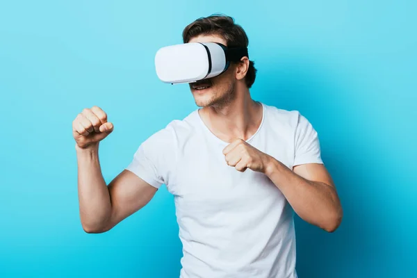 Man in white t-shirt and vr headset showing fists on blue background — Stock Photo