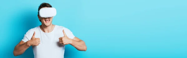 Panoramic shot of man showing thumbs up while using vr headset on blue background — Stock Photo