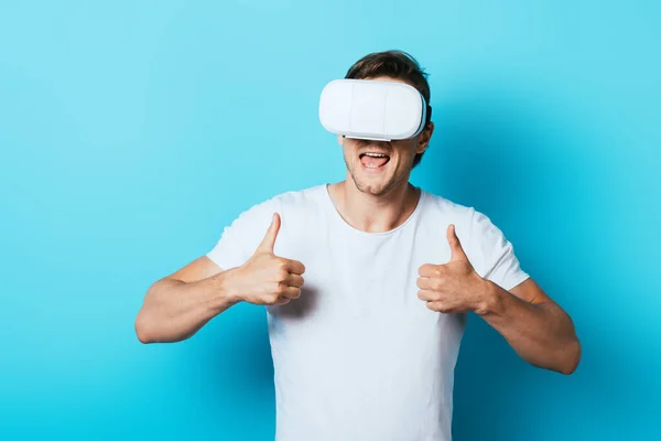 Junger Mann zeigt Daumen nach oben, während er Virtual-Reality-Headset auf blauem Hintergrund benutzt — Stockfoto