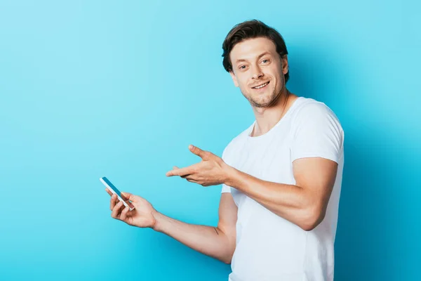 Молодий чоловік у білій футболці, спрямованій рукою на смартфон на синьому фоні — стокове фото