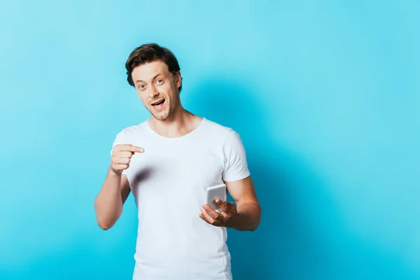 Hombre en camiseta blanca apuntando con el dedo al teléfono inteligente sobre fondo azul - foto de stock