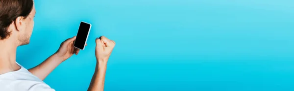 Panoramic shot of young man showing yes gesture while holding smartphone with blank screen on blue background — Stock Photo