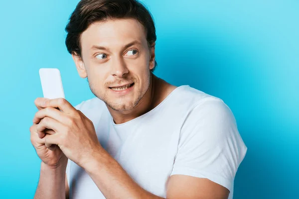 Hombre complicado sosteniendo el teléfono inteligente y mirando hacia otro lado sobre fondo azul - foto de stock