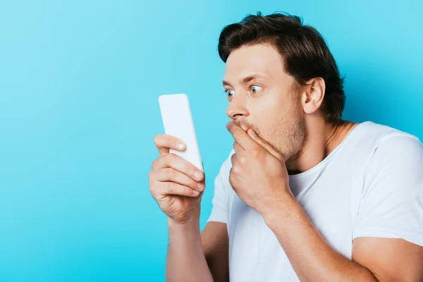 Shocked man covering mouth with hand while using smartphone on blue background — Stock Photo