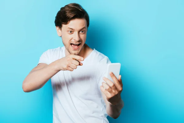 Foco seletivo do homem animado apontando com o dedo para o smartphone no fundo azul — Fotografia de Stock