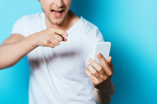 Ausgeschnittene Ansicht eines aufgeregten Mannes, der mit dem Finger auf das Smartphone auf blauem Hintergrund zeigt — Stockfoto