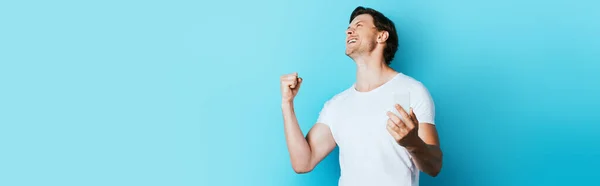 Panoramic shot of man showing yeah gesture and holding smartphone on blue background — Stock Photo