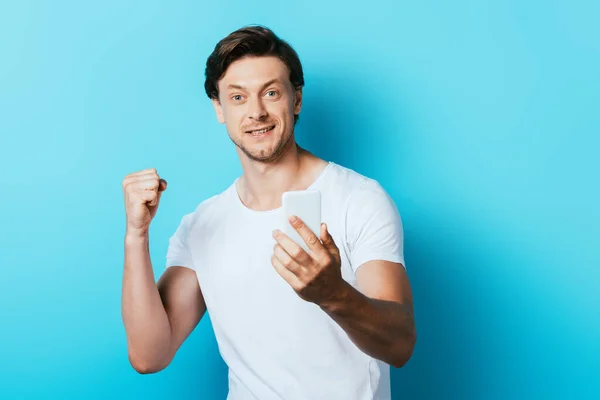 Young man looking at camera and showing yeah gesture while holding smartphone on blue background — Stock Photo