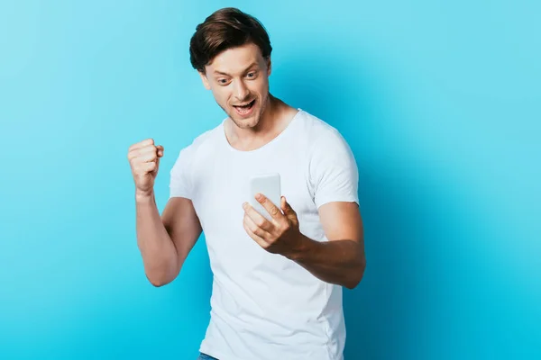 Man looking at smartphone and showing yeah gesture on blue background — Stock Photo