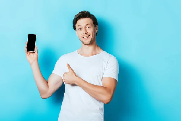 Young man showing thumb up and smartphone with blank screen on blue background — Stock Photo