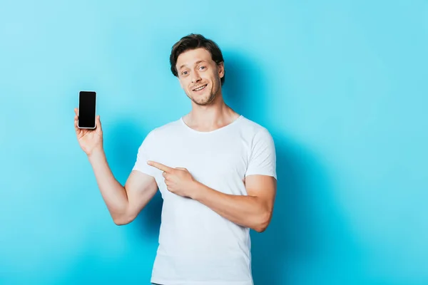 Mann im weißen T-Shirt zeigt auf Smartphone auf blauem Hintergrund — Stockfoto