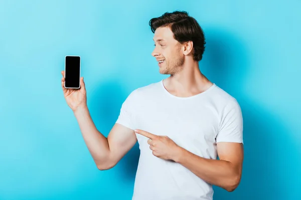 Mann schaut und zeigt mit dem Finger auf Smartphone mit leerem Bildschirm auf blauem Hintergrund — Stockfoto