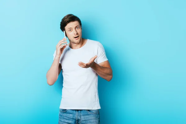 Joven apuntando con la mano mientras habla en el teléfono inteligente sobre fondo azul - foto de stock