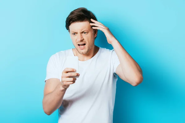 Angry man looking at smartphone on blue background — Stock Photo