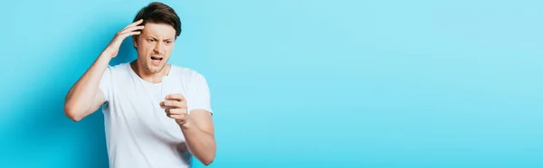 Panoramic shot of aggressive man looking at smartphone on blue background — Stock Photo
