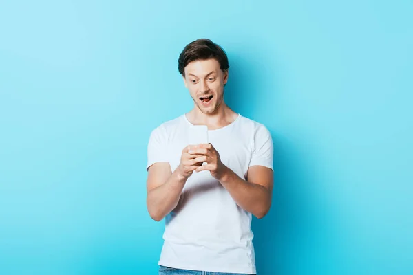 Homem animado em t-shirt branca usando smartphone no fundo azul — Fotografia de Stock