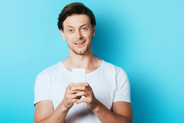 Young man looking at camera while using smartphone on blue background — Stock Photo