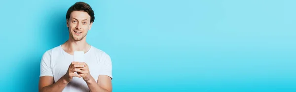 Panoramic shot of man looking at camera while using smartphone on blue background — Stock Photo