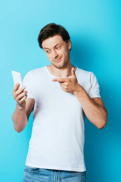 Hombre en camiseta blanca apuntando con el dedo al teléfono inteligente sobre fondo azul - foto de stock