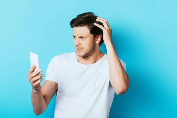 Hombre reflexivo con la mano cerca de la cabeza utilizando el teléfono inteligente sobre fondo azul - foto de stock