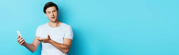 Panoramic shot of confused man looking at camera while pointing at smartphone on blue background — Stock Photo