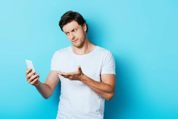 Hombre serio señalando con la mano en el teléfono inteligente sobre fondo azul - foto de stock